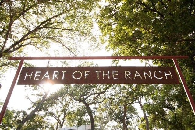 Rusted steel Heart of the Ranch sign hung from a red iron beam, backlit by the sun through the trees